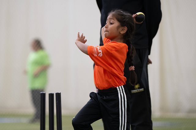 A young girl in an orange t-shirt is throwing a cricket ball.