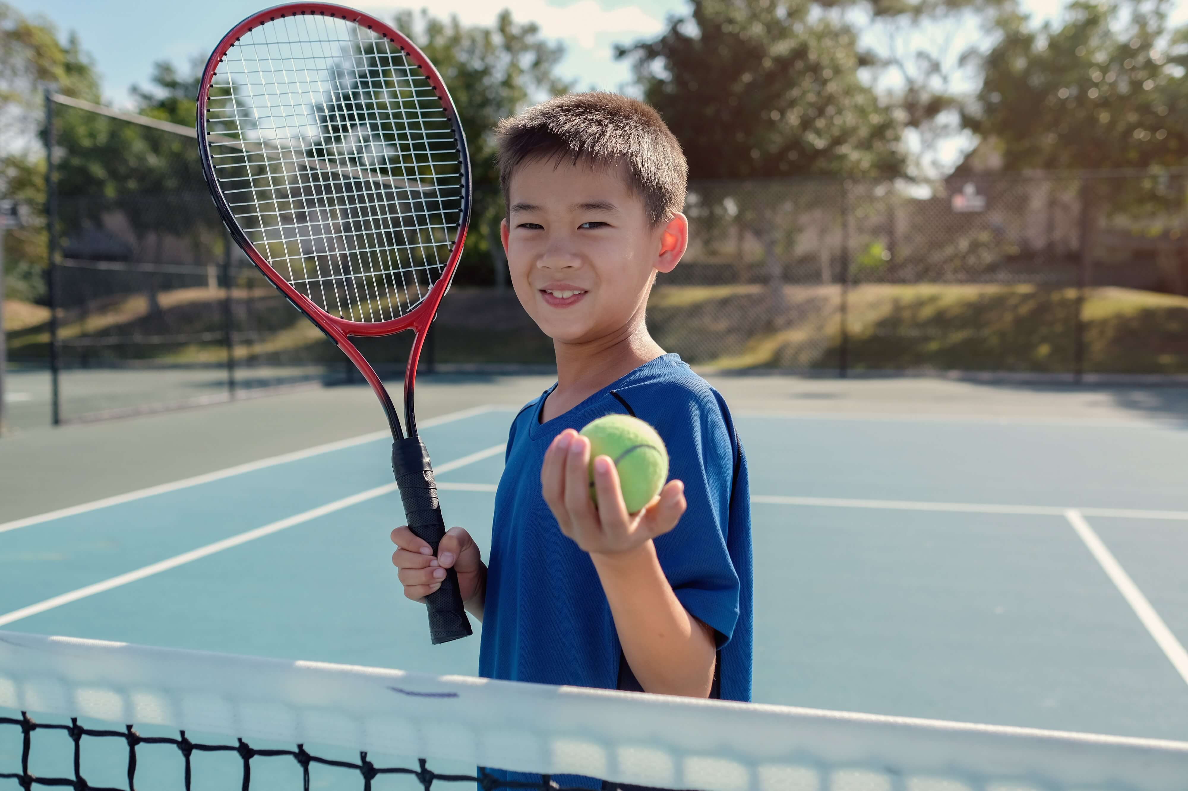 Boy on blue outdoor tennis court holding a tennis racket in his right hand and a tennis ball in his left hand
