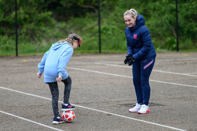 A happy football coach clapping hands looks on as a young girl focuses on controlling the ball with her foot in the playground