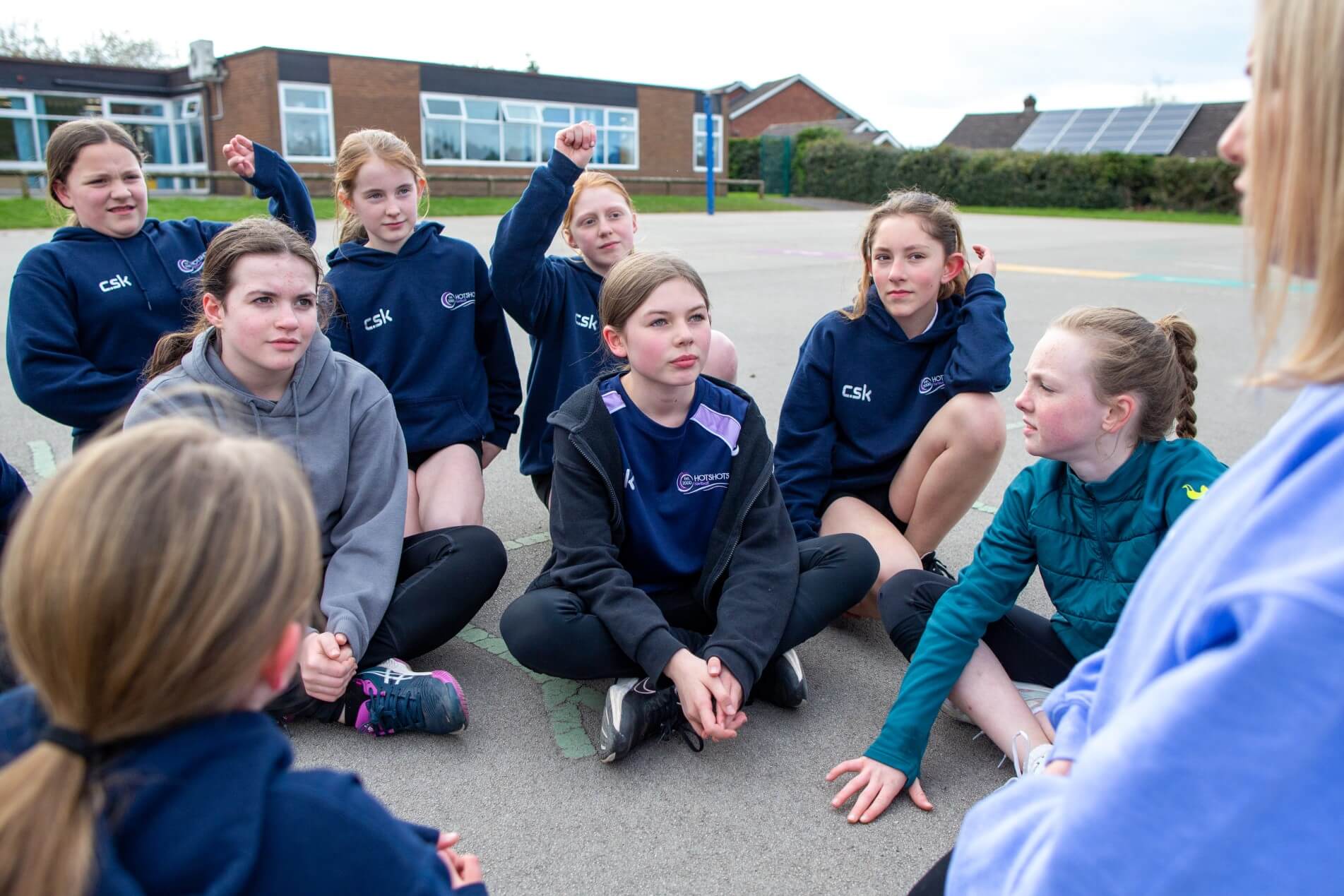 A group of young girls are sat on the playground talking to their female coach. 