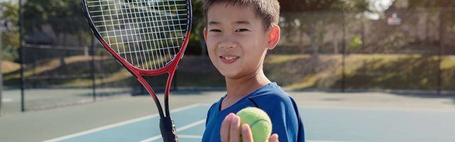 Young boy wearing a bright blue t-shirt is holding a tennis ball to camera and has a racket in his right hand