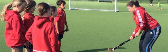 Female hockey coach is holding a hockey stick demonstrating technique to a group of young girls
