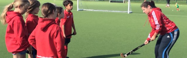 Female hockey coach is holding a hockey stick demonstrating technique to a group of young girls