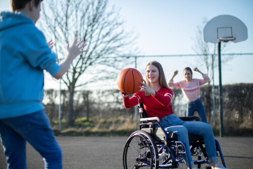 Teen in a red top and in a wheelchair is about to pass a basketball at a person in a blue top