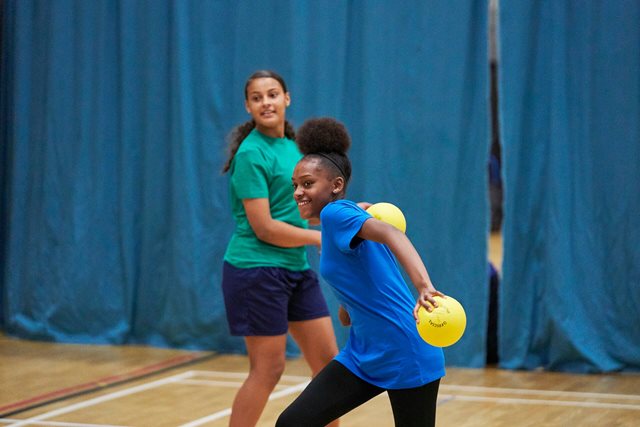 A teenager throws a dodgeball with enthusiasm in a sports hall, smiling with excitement as they aim for the target