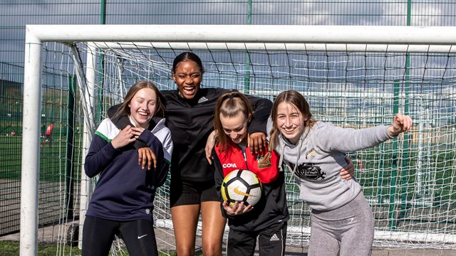 Four girls standing together in front of a football net with a ball celebrating a goal