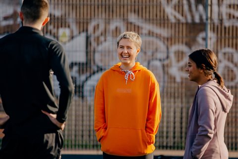 Two young people, one wearing an orange jumper and one a grey jumper, smile at their coach and in relaxed mood with their hands in the pockets of their jumpers