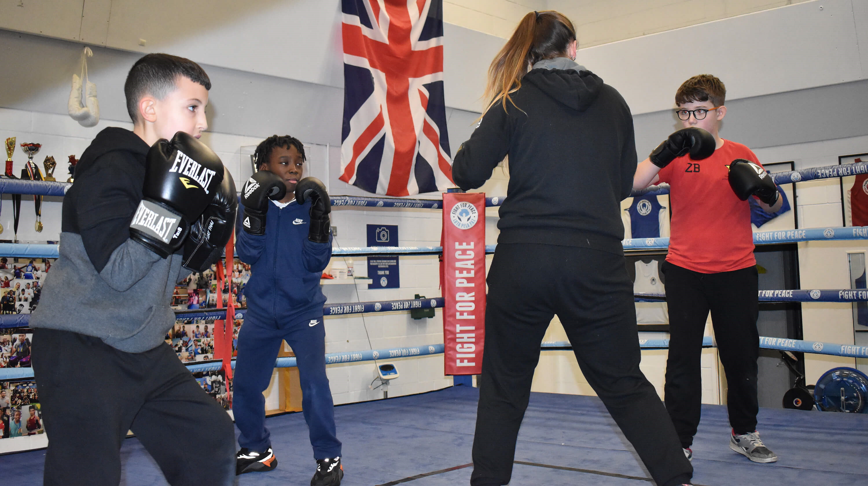 A coach stands in the ring surrounded by three young boxers who are practising their stance and footwork