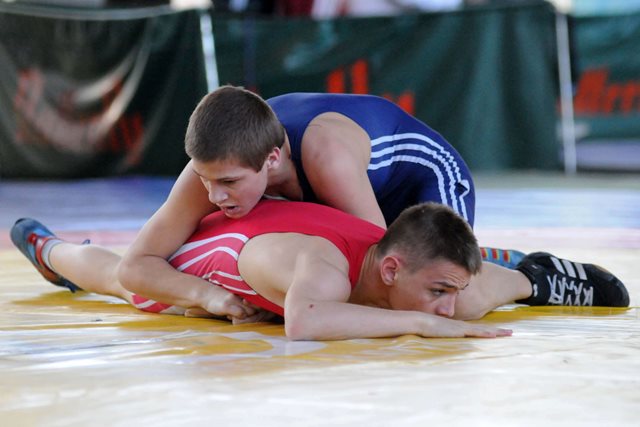 Two young boys, one in a blue singlet and the other in a red singlet are on the ground in a wrestling hold