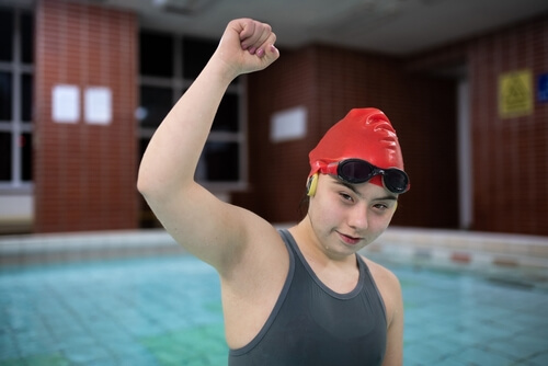 Disabled girl in red swimming cap and grey swimming costume is air punching as she is stood by a swimming pool. 