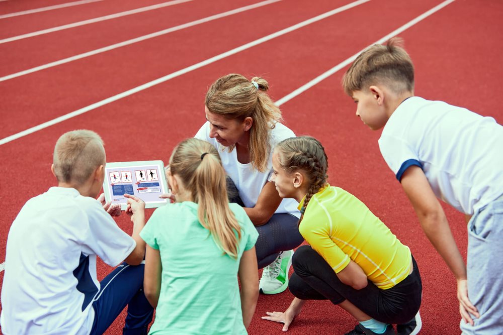 Excited children huddle around their athletic coach as she displays a fun activity on her iPad