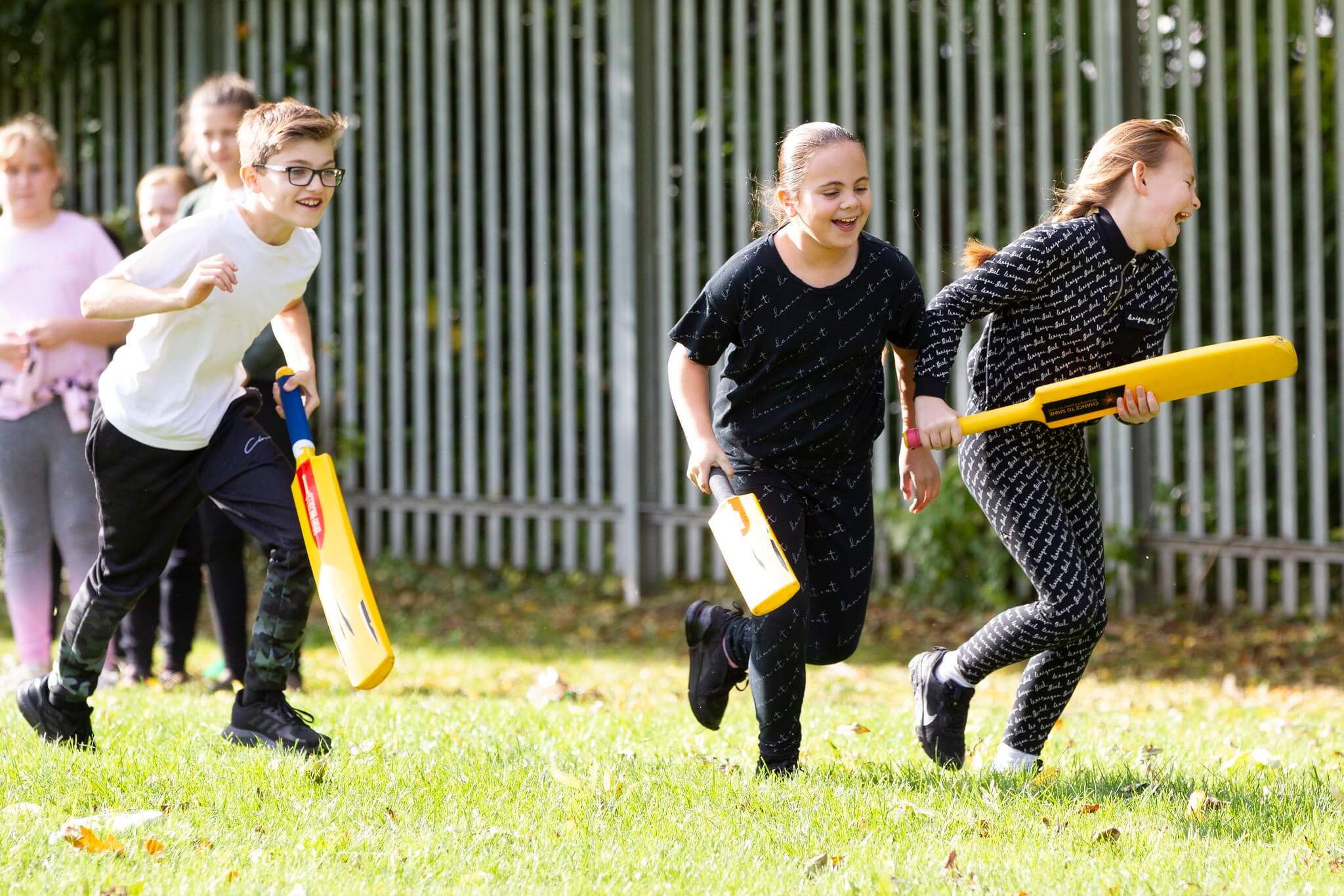 Three children sprint forwards holding their cricket bats on a school sports field