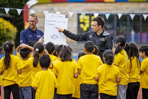 A coach/teacher outlines a cricket game watched by a group of school children in yellow sports tops