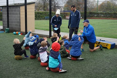 A group of very small children are sat on the ground whilst their coaches talk to them 