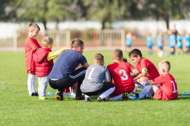 A male coach in a blue t-shirt is kneeling on the ground chatting to a group of boys in red football kits who are sat on the ground. 