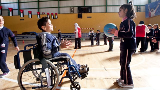 A young girl about to throw a ball to a young boy in a wheelchair in an indoor sports hall