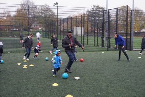 small child in blue coat dribbles between cones on outdoor football pitch closely watched by coach