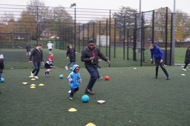 small child in blue coat dribbles between cones on outdoor football pitch closely watched by coach