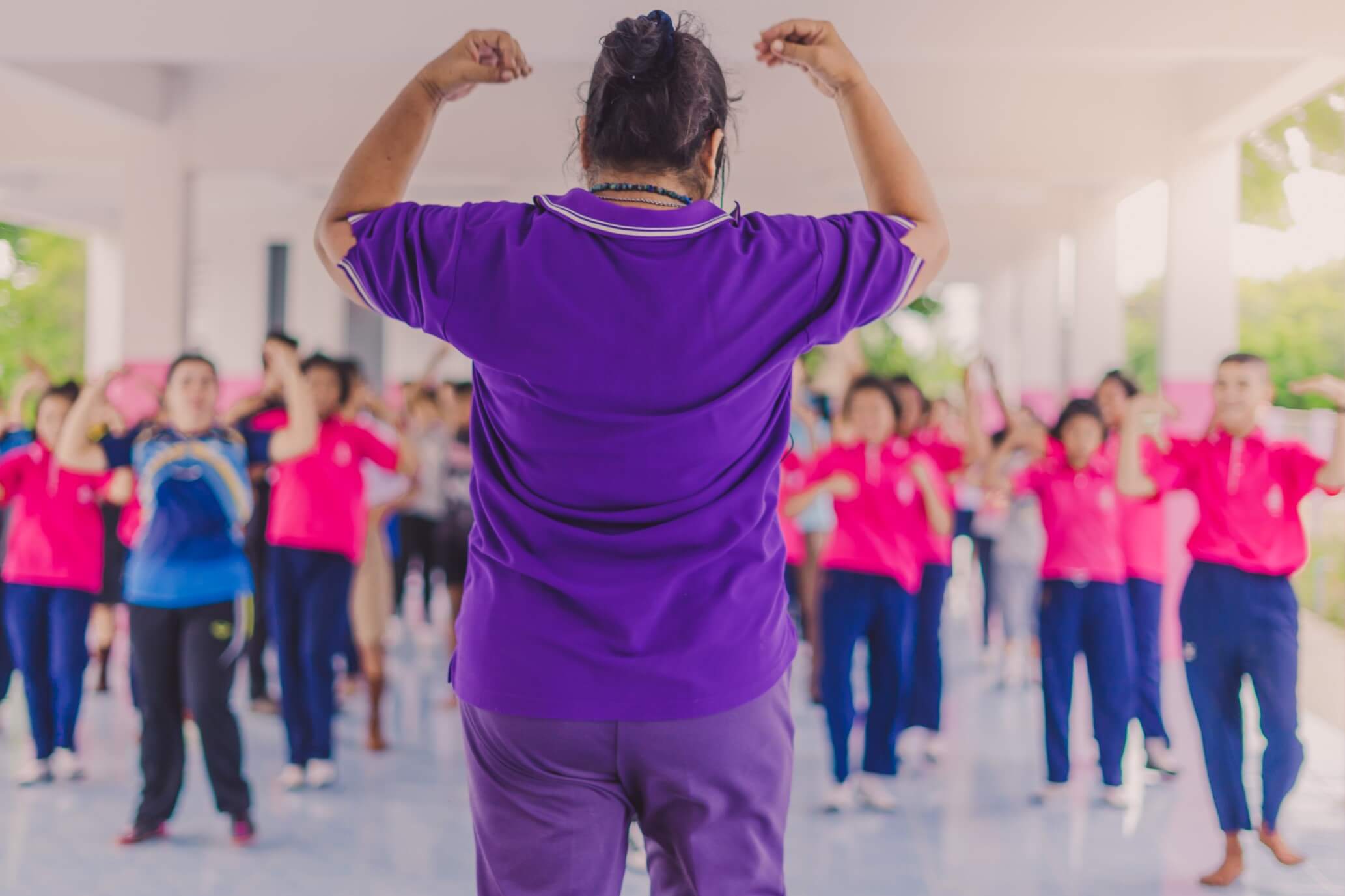 Woman coach in a purple t-shirt is stood in front of a class of children wearing pink t-shirts directing them in a warm-up activity.