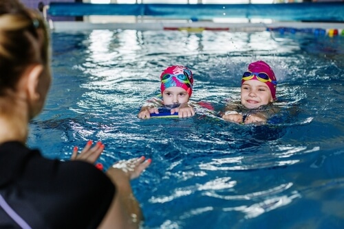 Two girls in the pool learning to swim by using a float with coach on the edge of the pool