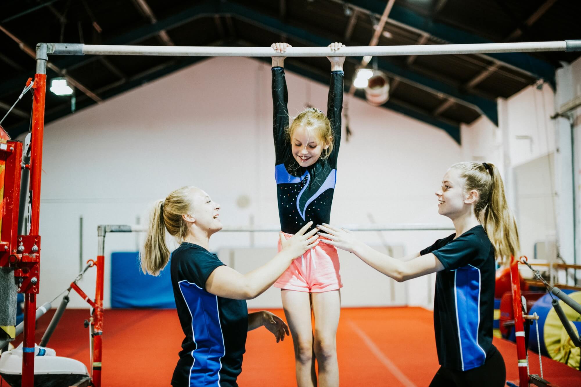 Two coaches stand either side of a young gymnast as she holds on to the bar apparatus
