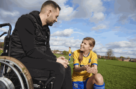 Male coach in wheelchair talks to a girl dressed in a yellow and blue football strip