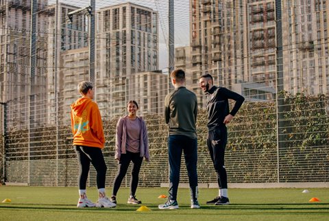 A coach and three young people standing on a fenced Astroturf pitch with tower blocks in the background
