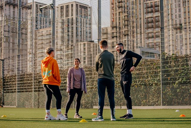A coach and three young people standing on a fenced Astroturf pitch with tower blocks in the background
