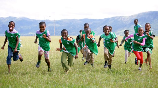 Image from Nigeria showing a smiling children in a line wearing green sports kit sprinting forwards